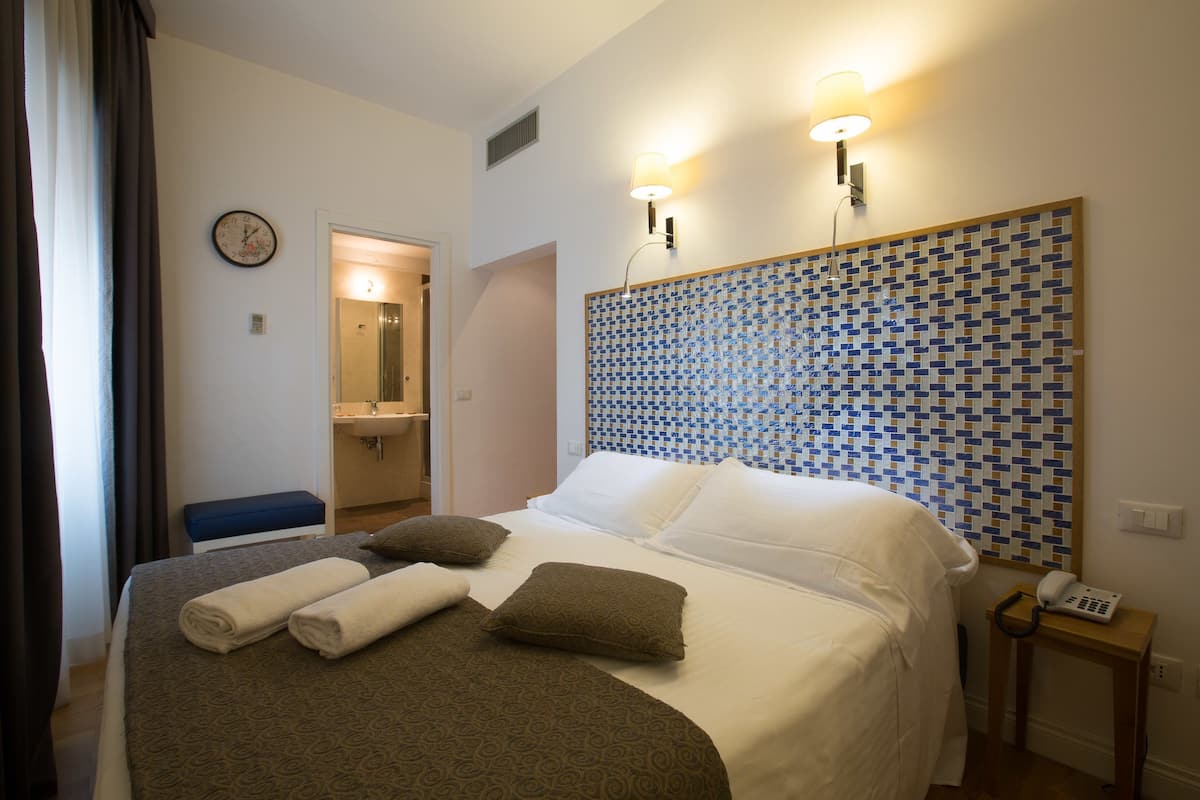 A hotel room with a bed, a bedside table, a clock, and a bathroom visible through an open door.