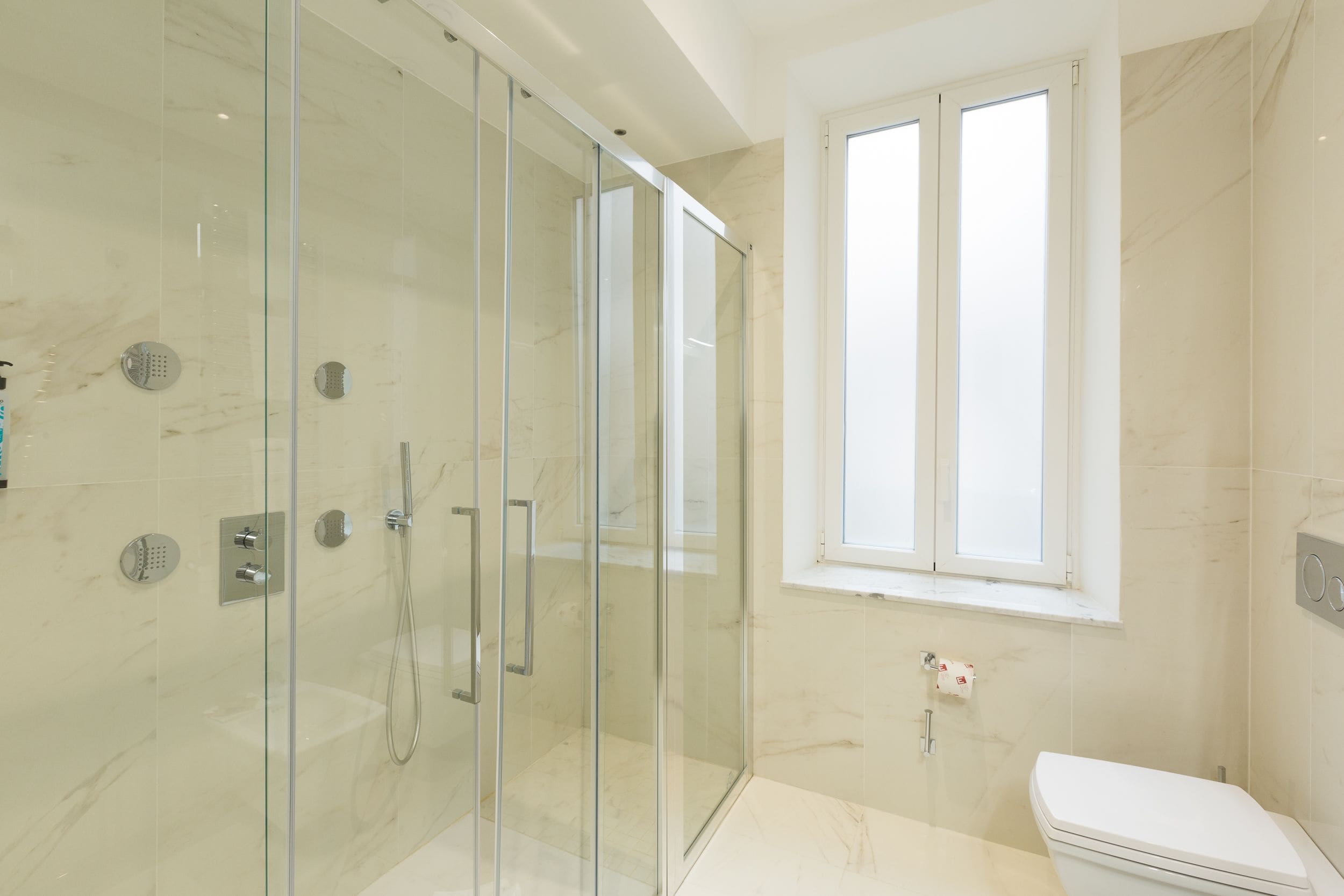 A modern bathroom with a glass-enclosed shower, a toilet, and a window providing natural light.