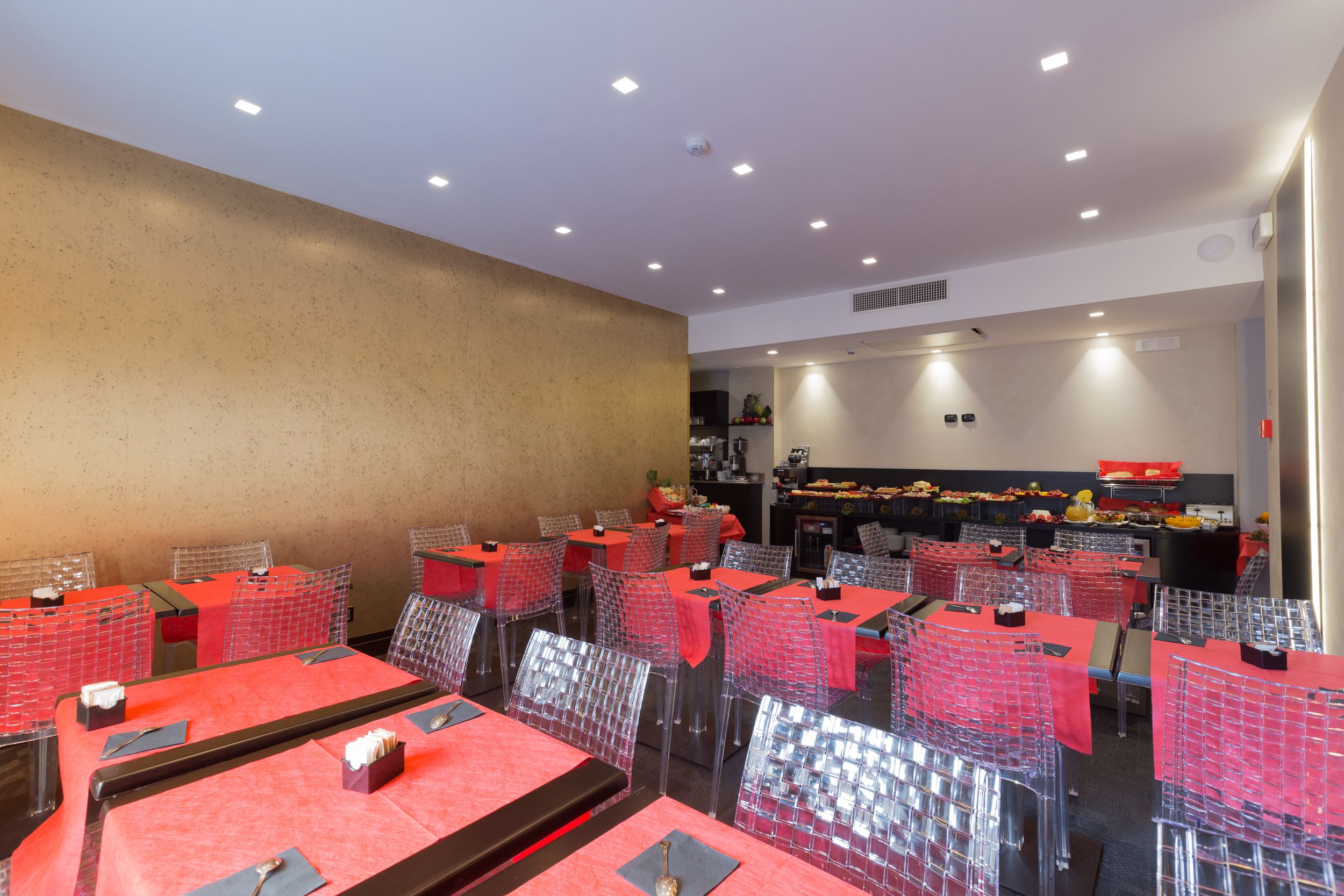 A restaurant with tables covered in red tablecloths and clear chairs, with a buffet area in the background.