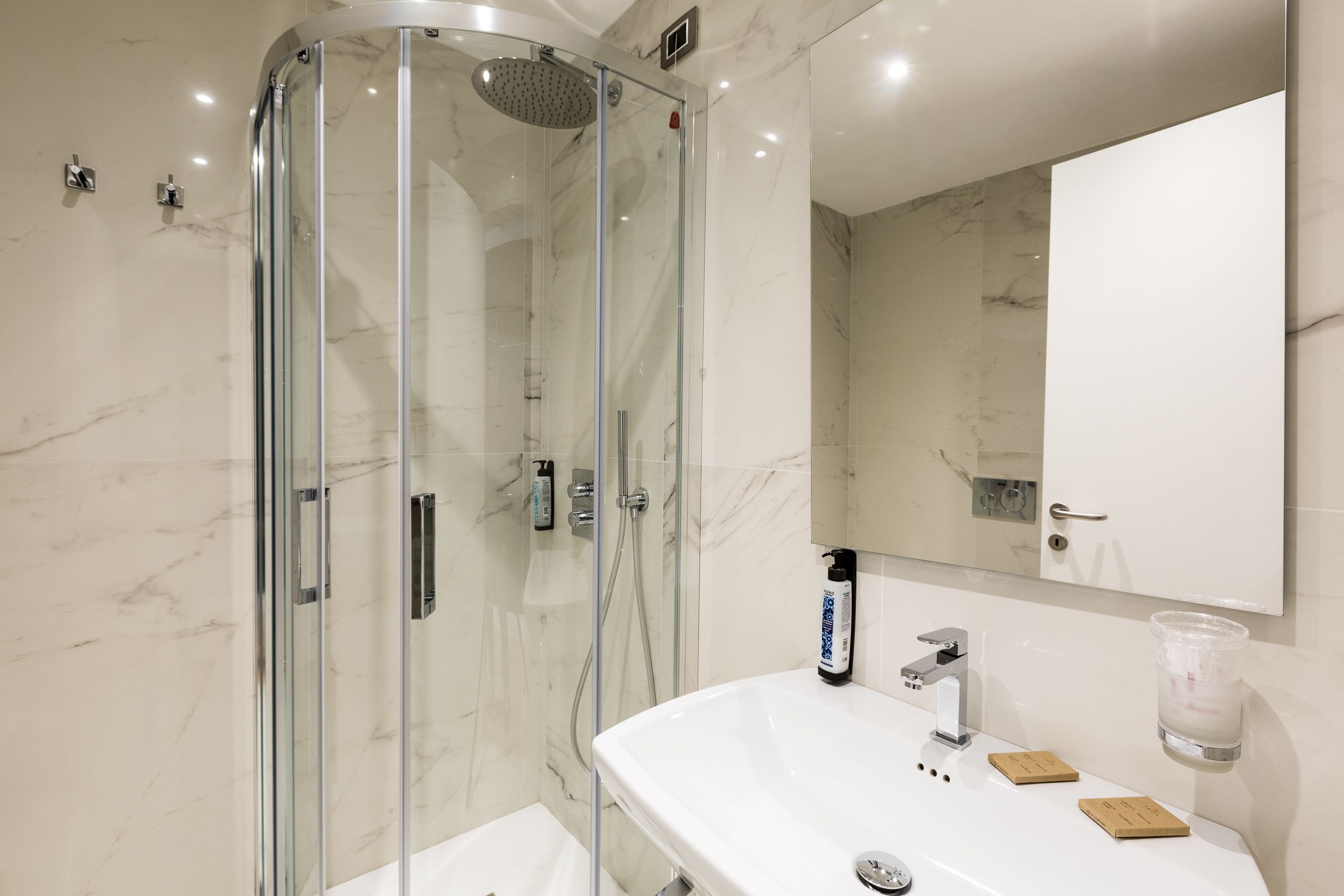 Un baño moderno con ducha de vidrio, un lavamanos blanco con jabón y un espejo sobre el lavamanos.