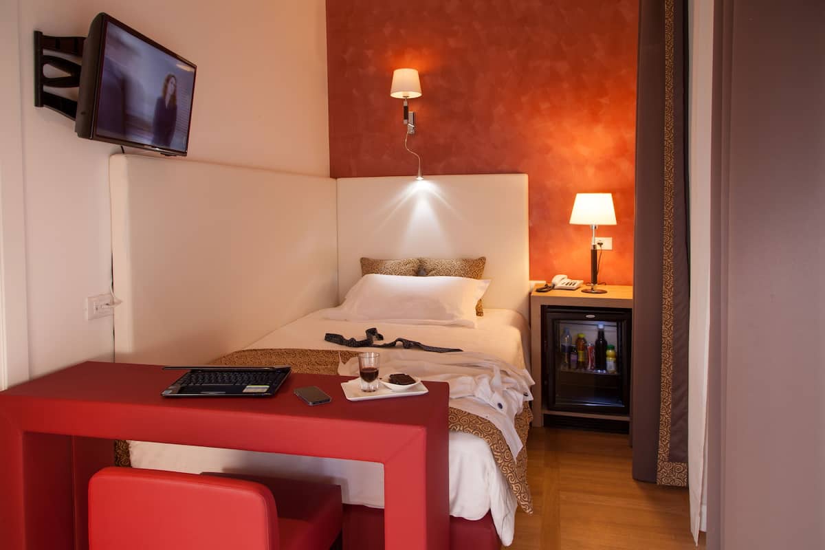 A hotel room with a bed, a desk with a laptop, a chair, a bedside table with a lamp, and a minibar.