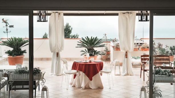 A dining area with a table set for four, surrounded by chairs, with a view of the sea through large glass doors. - B&B Triskéles (Agrigento)