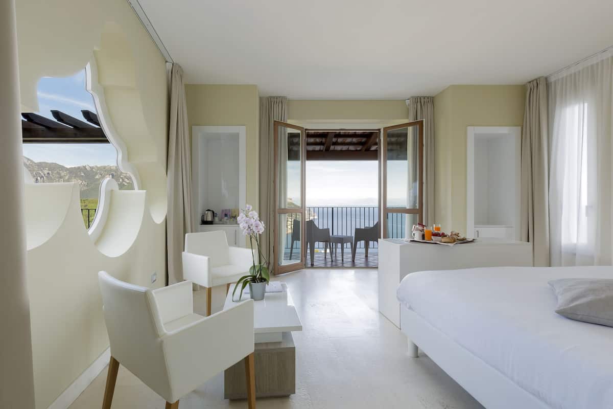 A hotel room with a bed, a desk, a chair, a small table with a potted plant, and a balcony with a view of the sea.