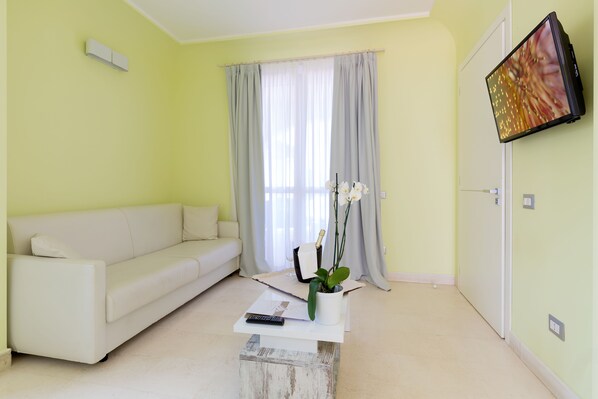 Un salón con un sofá blanco, una mesa de centro con un control remoto, una planta en maceta y un televisor montado en la pared.