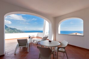 Un balcón con sillas y una mesa blancos, con vistas panorámicas al mar y las montañas.