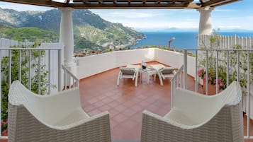 A balcony with wicker chairs and a table, overlooking a mountainous landscape and the sea.