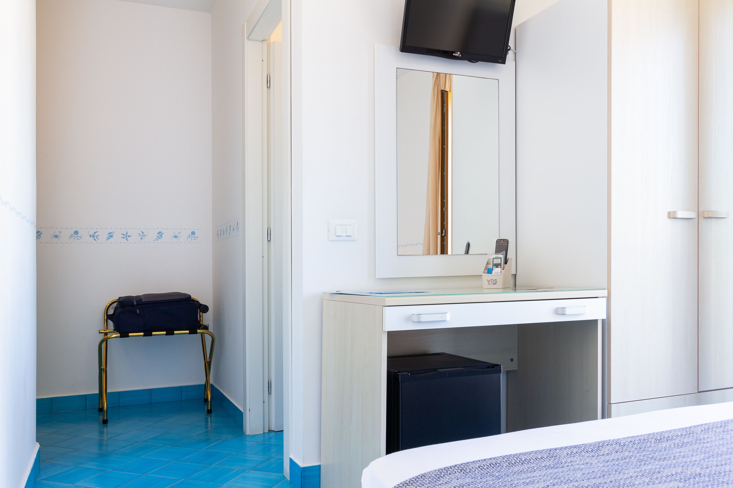 Habitación de hotel con cama, escritorio con espejo, taburete pequeño y cubo de basura.
