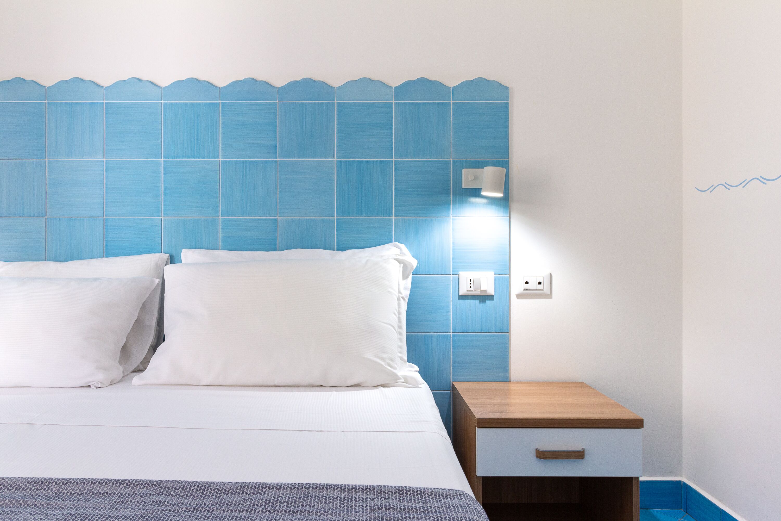 A neatly made bed with white linens and pillows, a wooden nightstand with a drawer, and a blue tiled headboard.