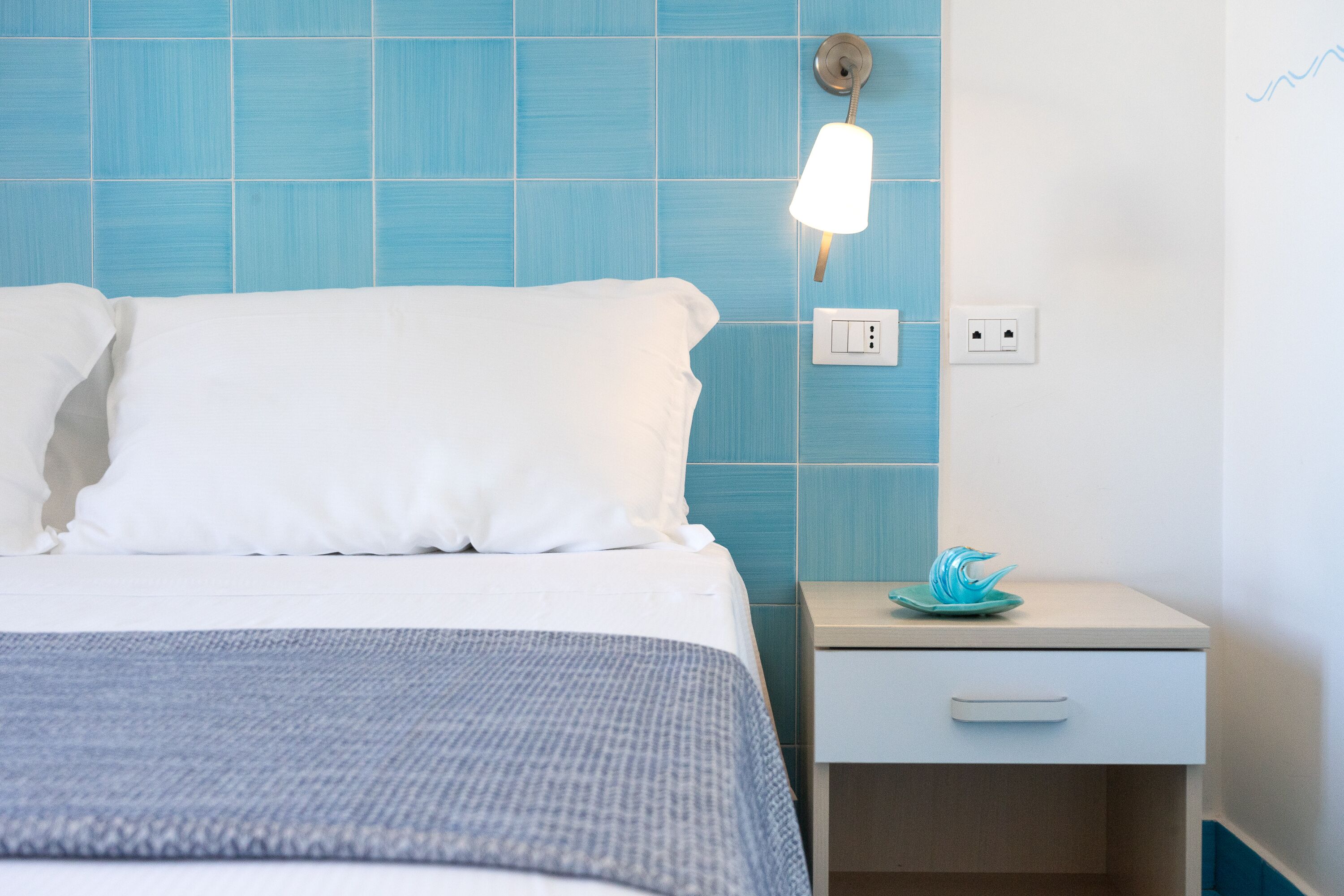 A neatly made bed with white linens and a blue patterned bedspread, a bedside table with a blue decorative object, and a wall with blue tiles and a wall-mounted light fixture.