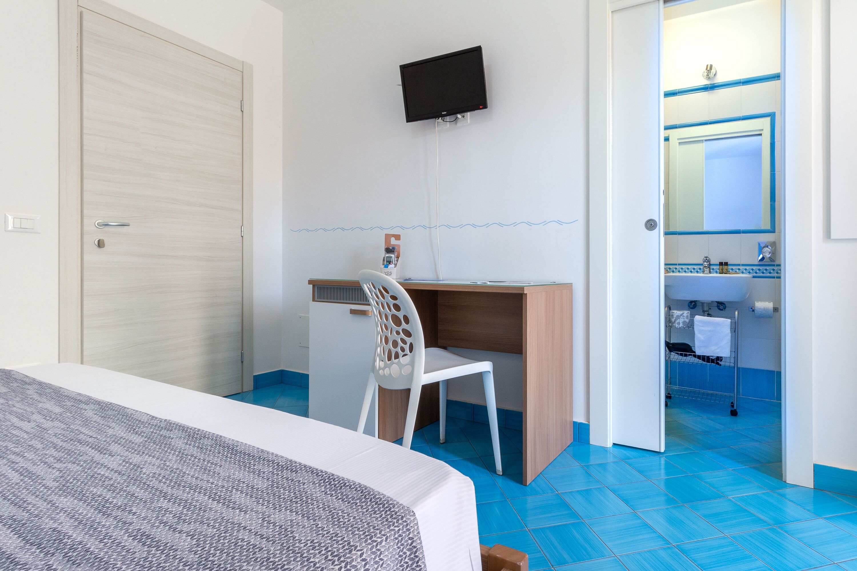 A hotel room with a bed, a desk with a chair, a television mounted on the wall, and a bathroom visible through an open door.