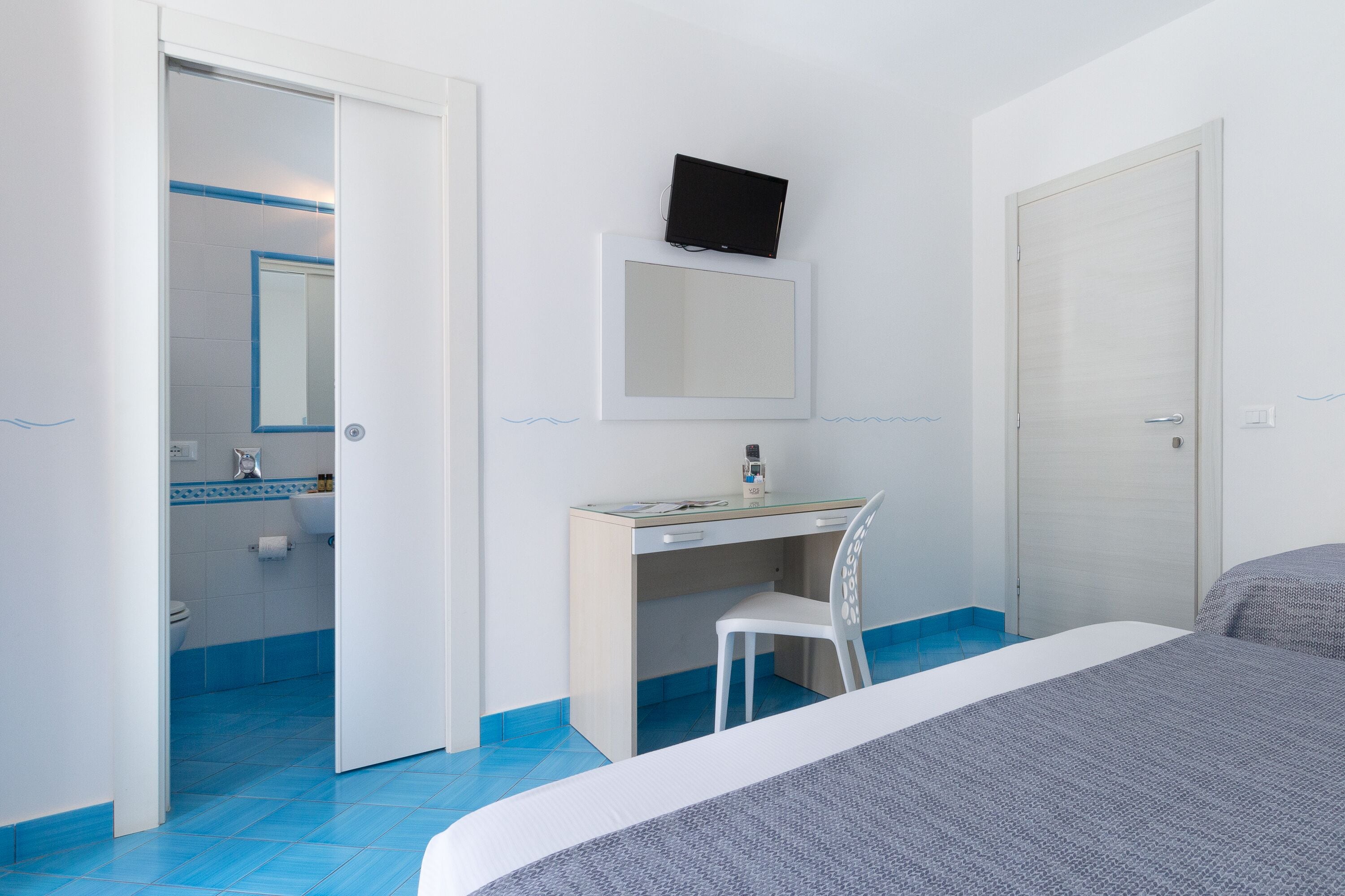 A bedroom with a bed, a desk with a chair, a television on the wall, and a bathroom visible through an open door.