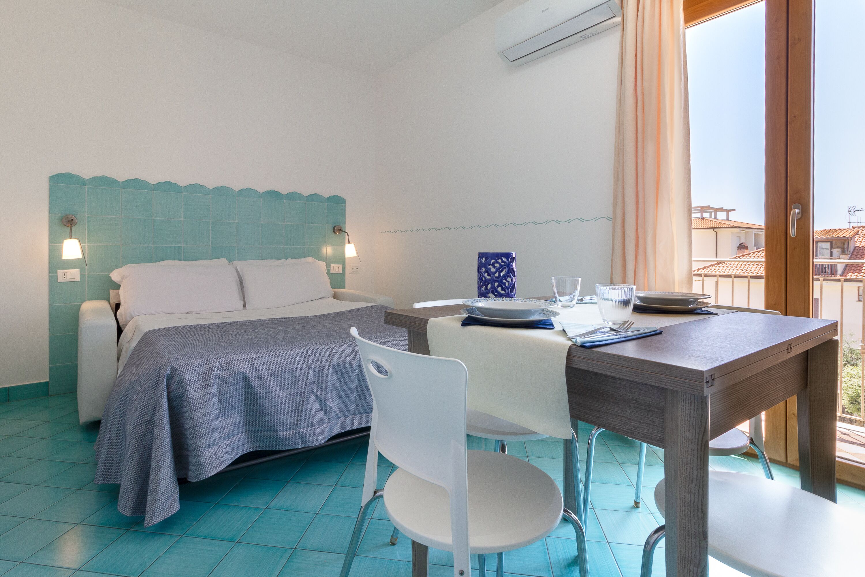A bedroom with a bed, a bedside table, a chair, a dining table, and a view of buildings through a window.