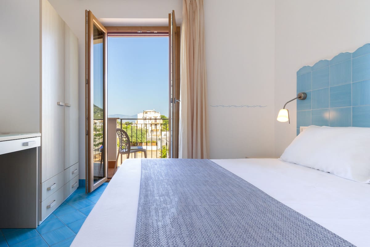 Ein Schlafzimmer mit Bett, Schreibtisch und einem Balkon mit Blick auf Gebäude und Grünflächen.