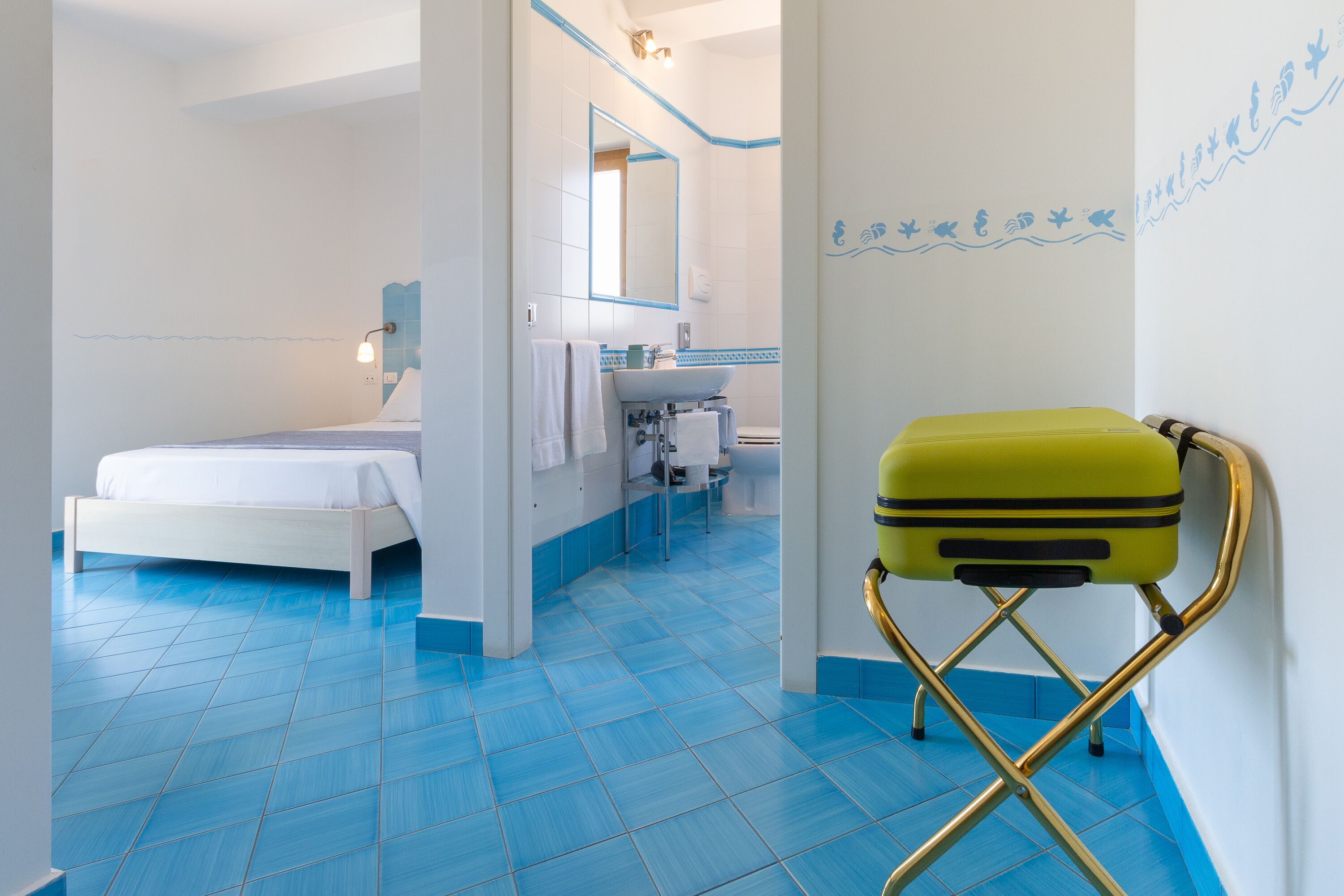Une chambre d’hôtel avec un lit, une salle de bain comprenant un lavabo et un miroir, et une valise posée sur une chaise.