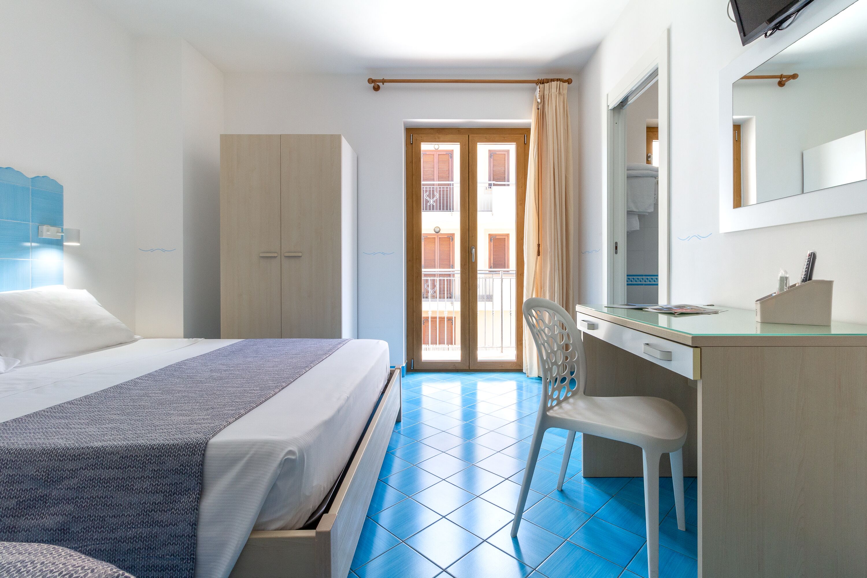A hotel room with a bed, a desk, a chair, a wardrobe, and a balcony with a view of the building.