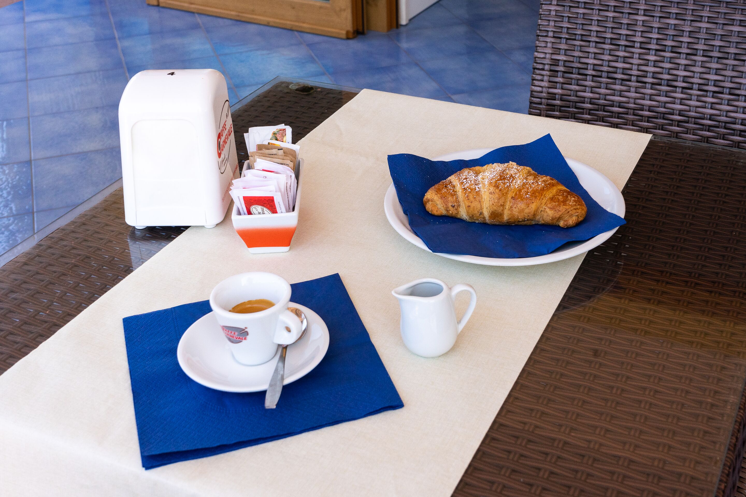 A table with a croissant, a cup of coffee, a small pitcher, and a tissue box.