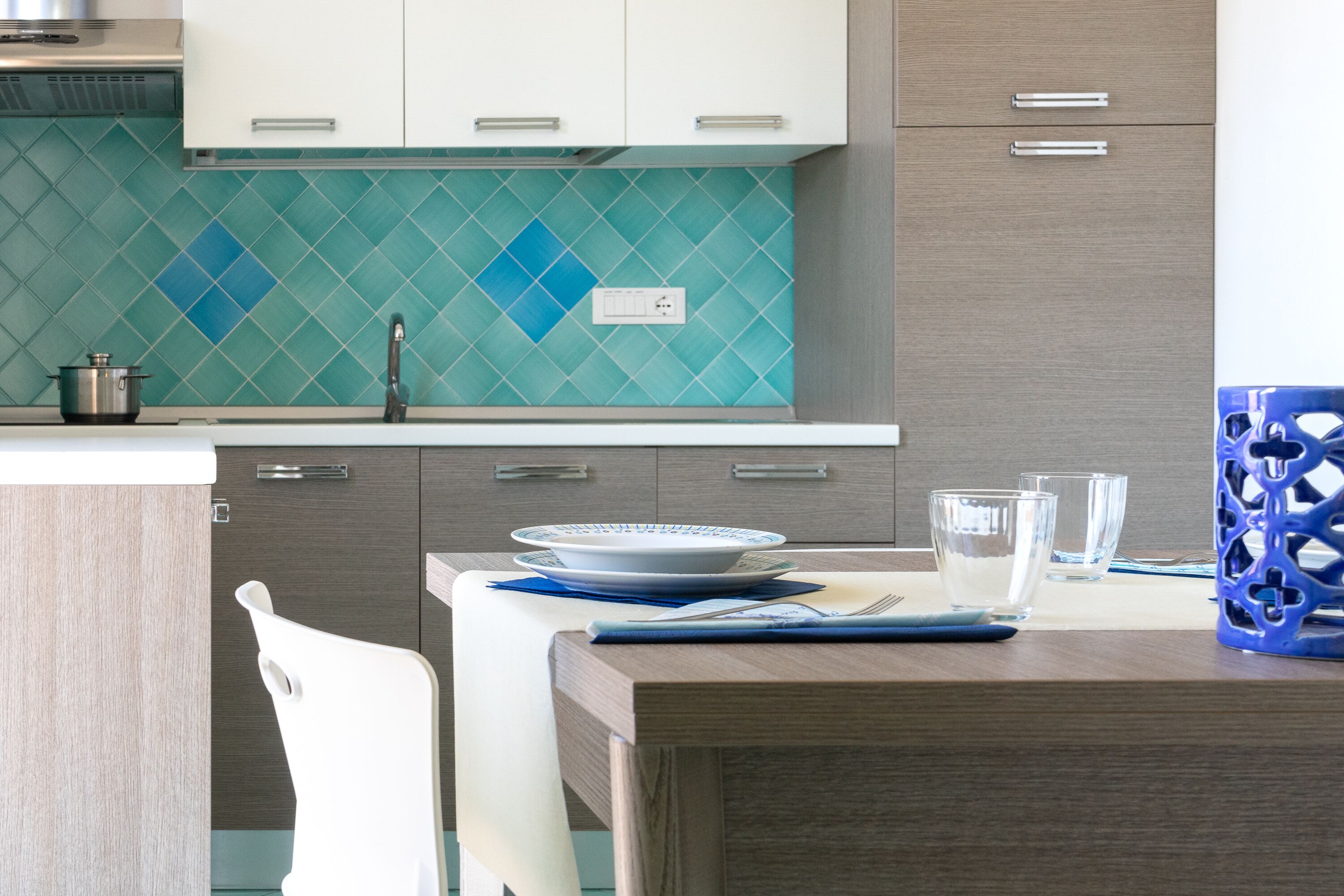 A modern kitchen with a teal backsplash, wooden cabinets, and a dining area with a white chair and a blue-patterned placemat.