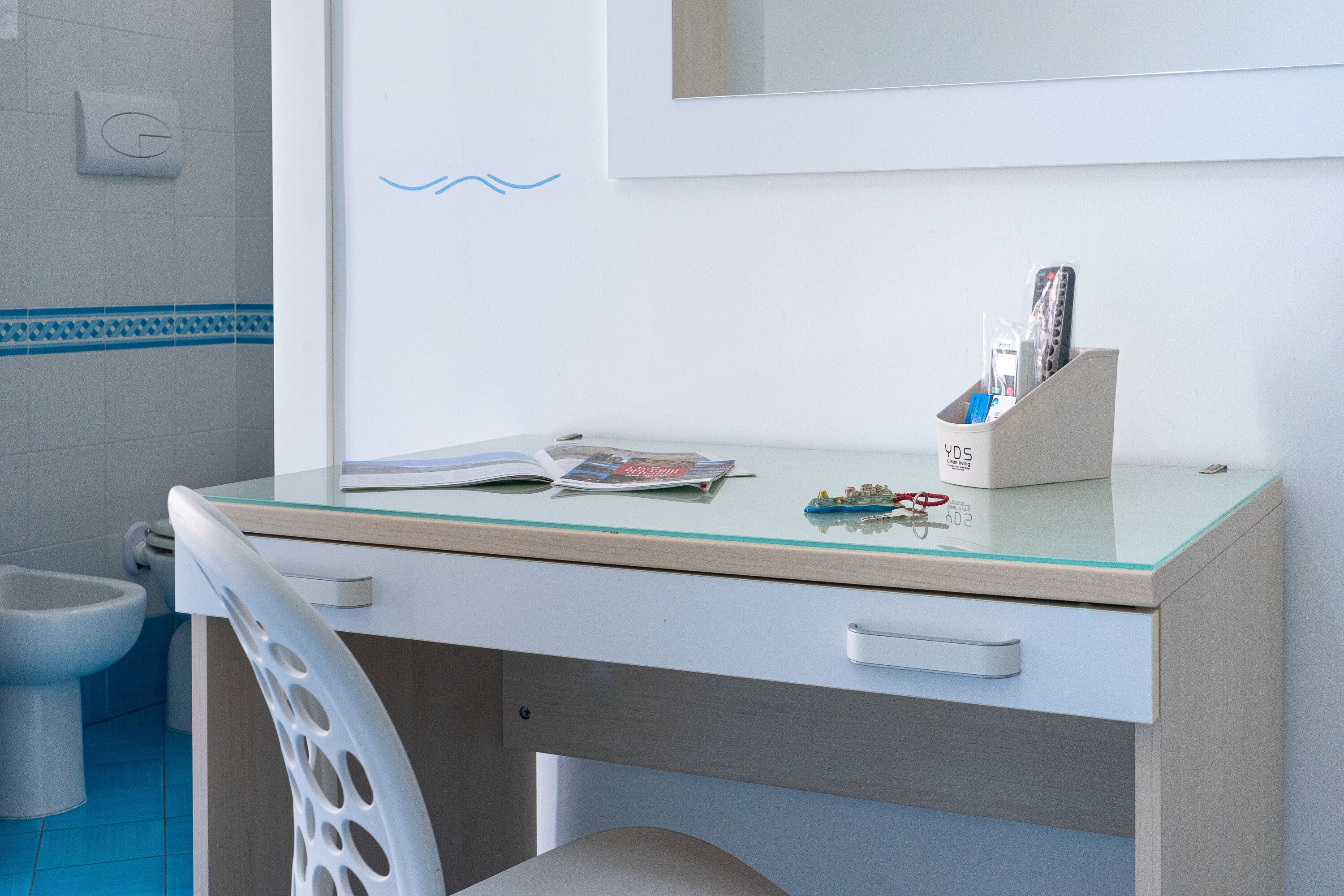 A bathroom with a white toilet, a bidet, and a glass-topped desk with a book and remote controls.