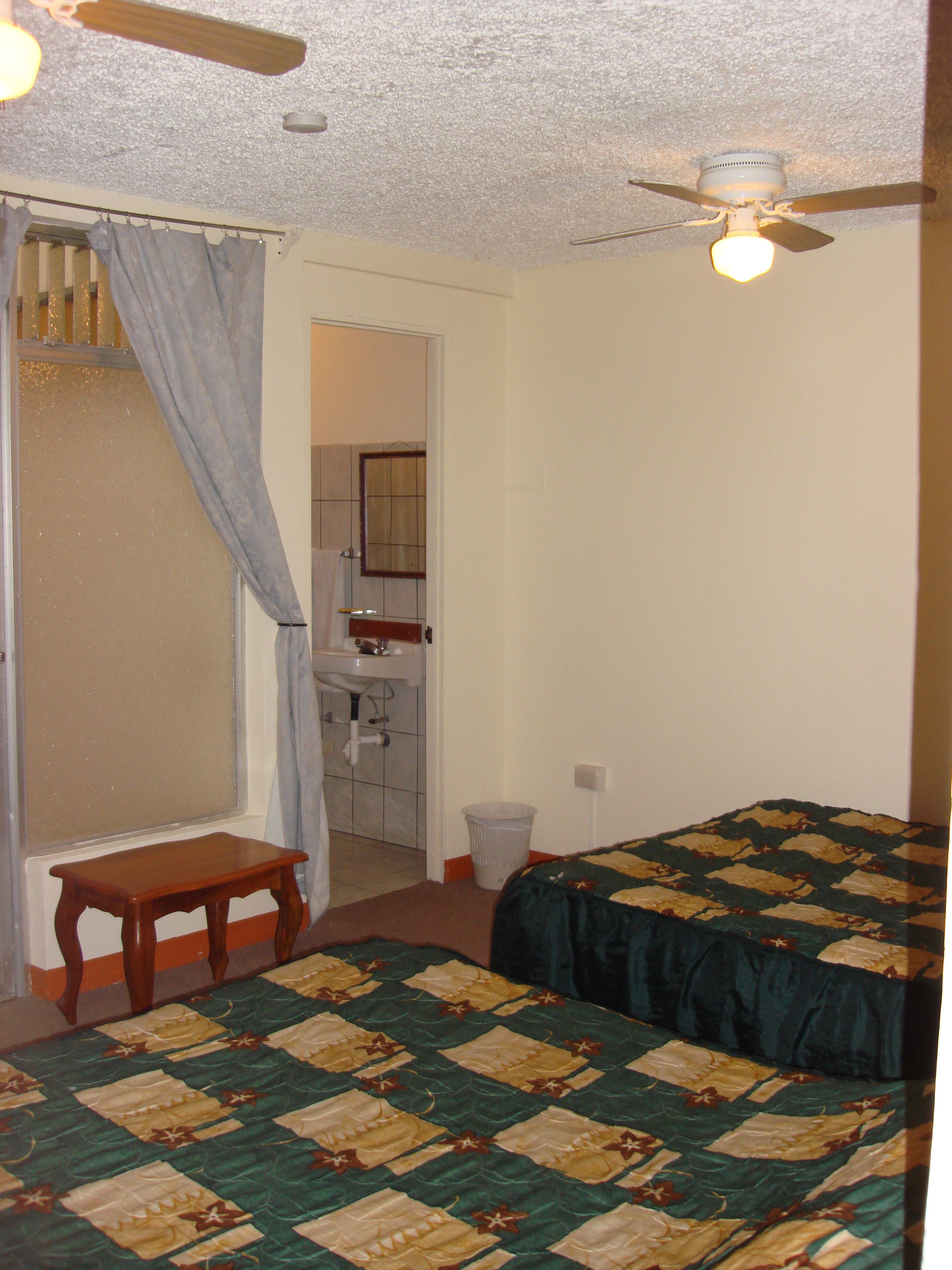 A bedroom with a bed, a small wooden bench, a ceiling fan, and a bathroom visible through an open door.