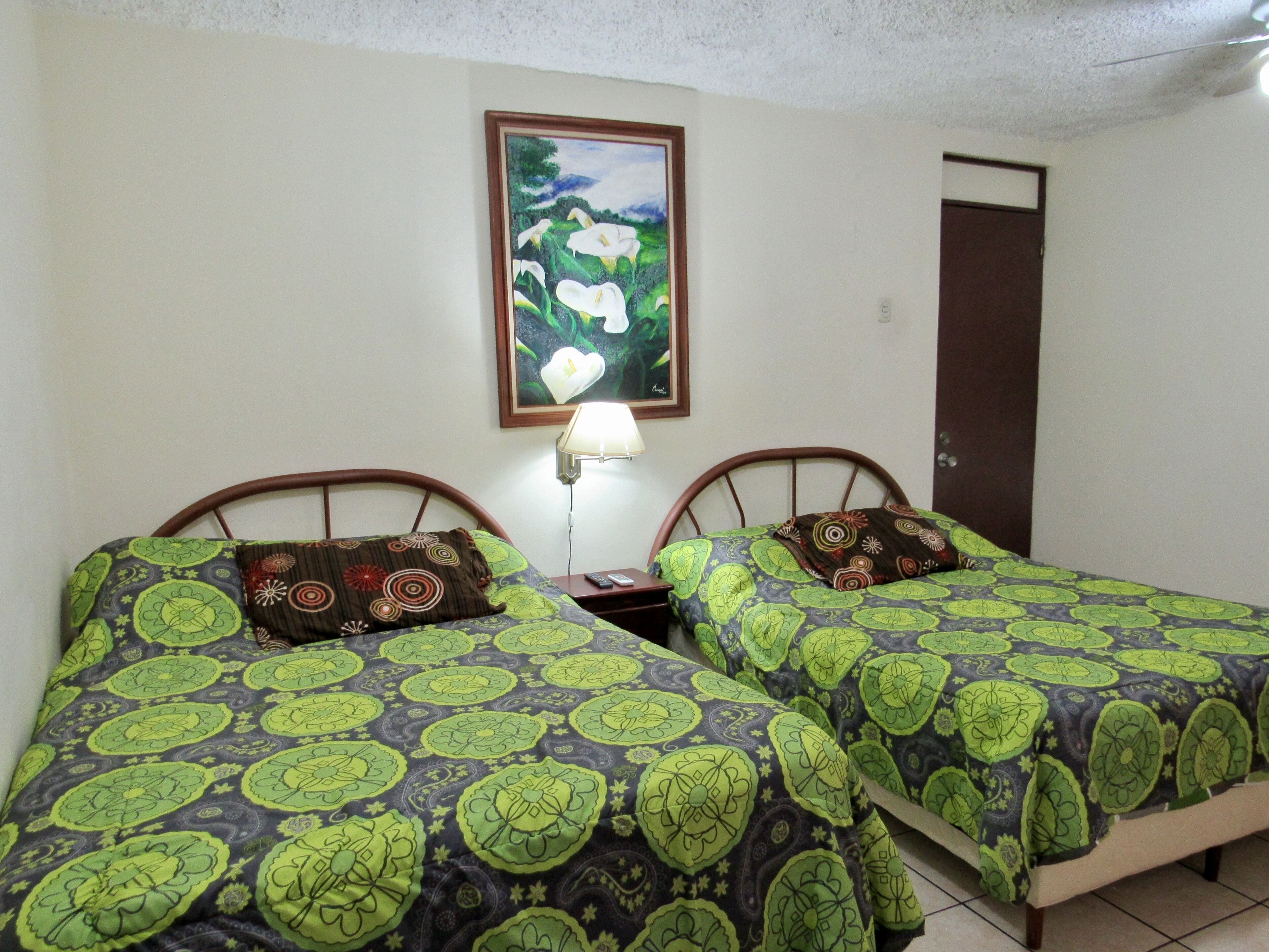 Una habitación con dos camas, cada una con ropa de cama estampada, una mesita de noche con lámpara y un cuadro enmarcado en la pared.