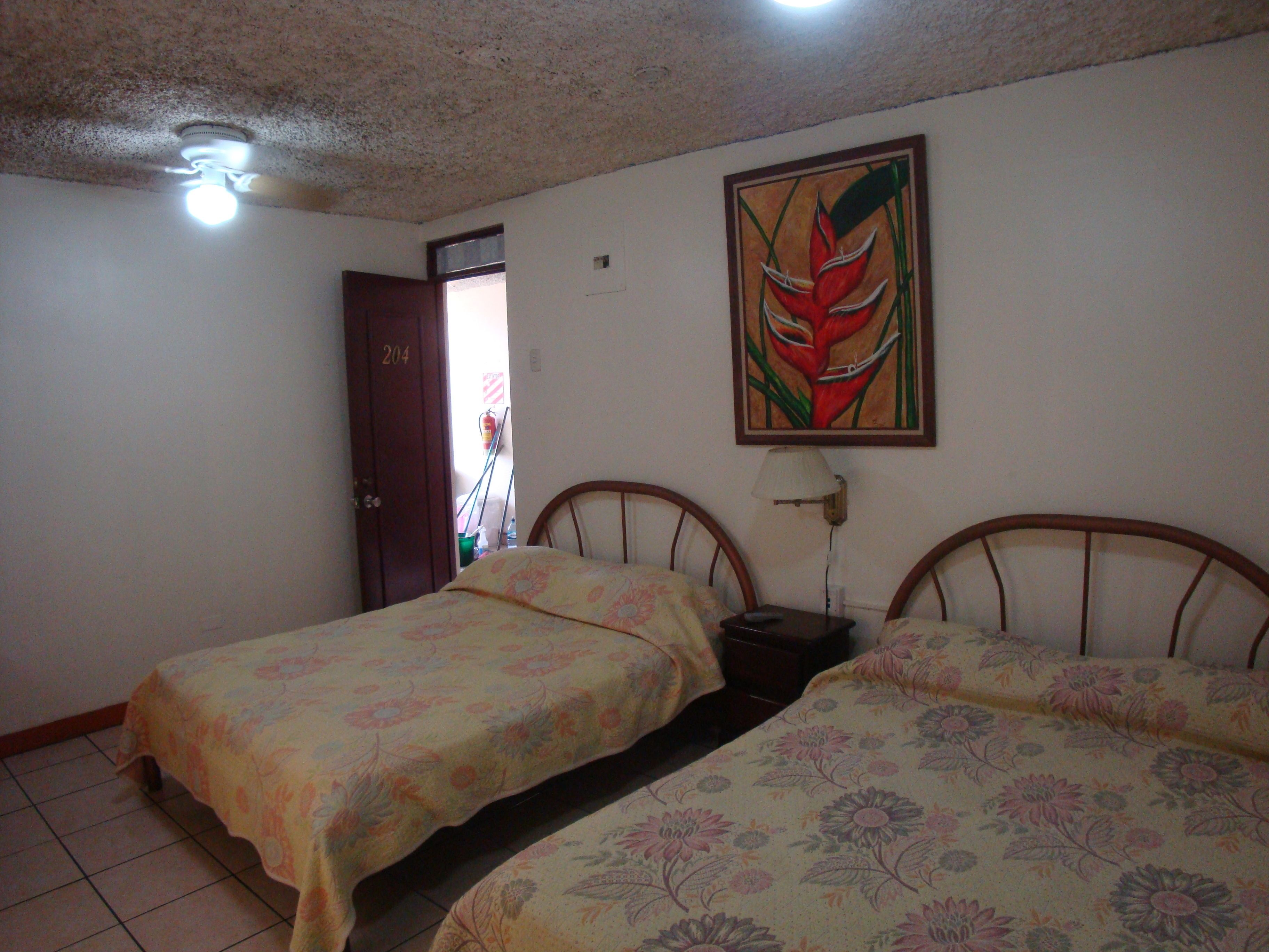 A room with two beds, a ceiling fan, a door, and a painting on the wall.
