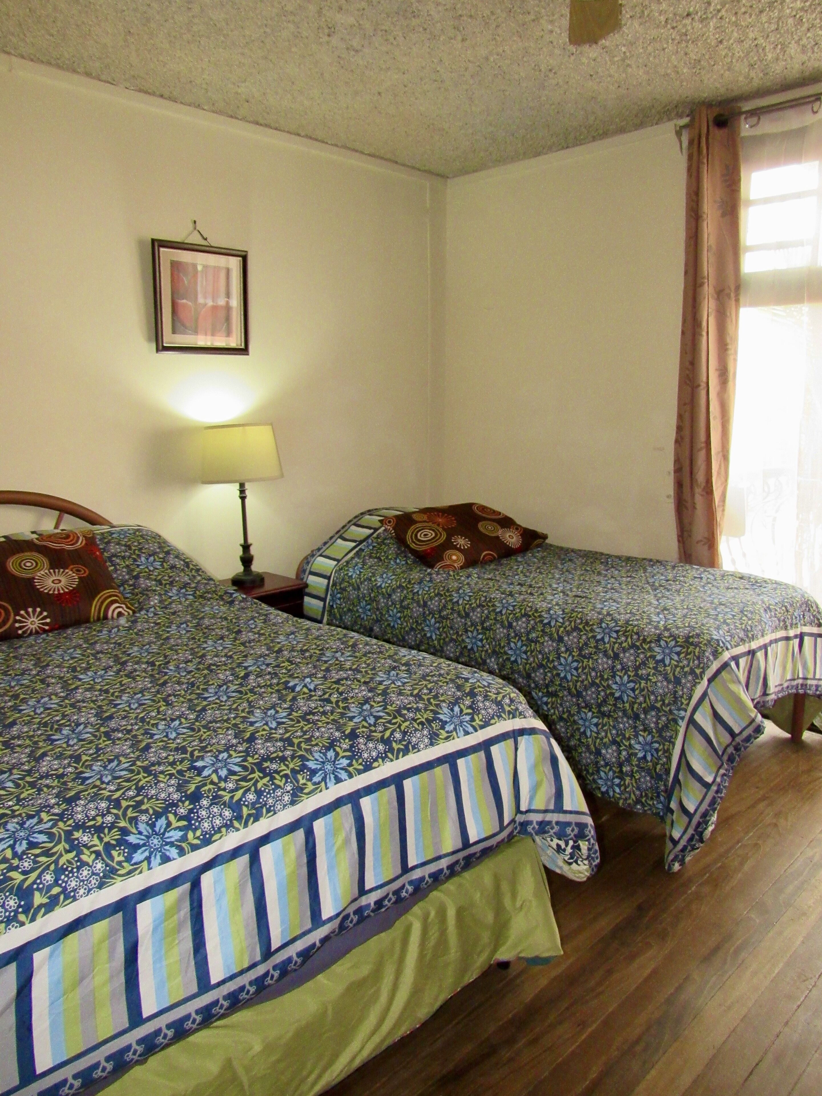 A room with two beds, a framed picture on the wall, a lamp, and a window with curtains.