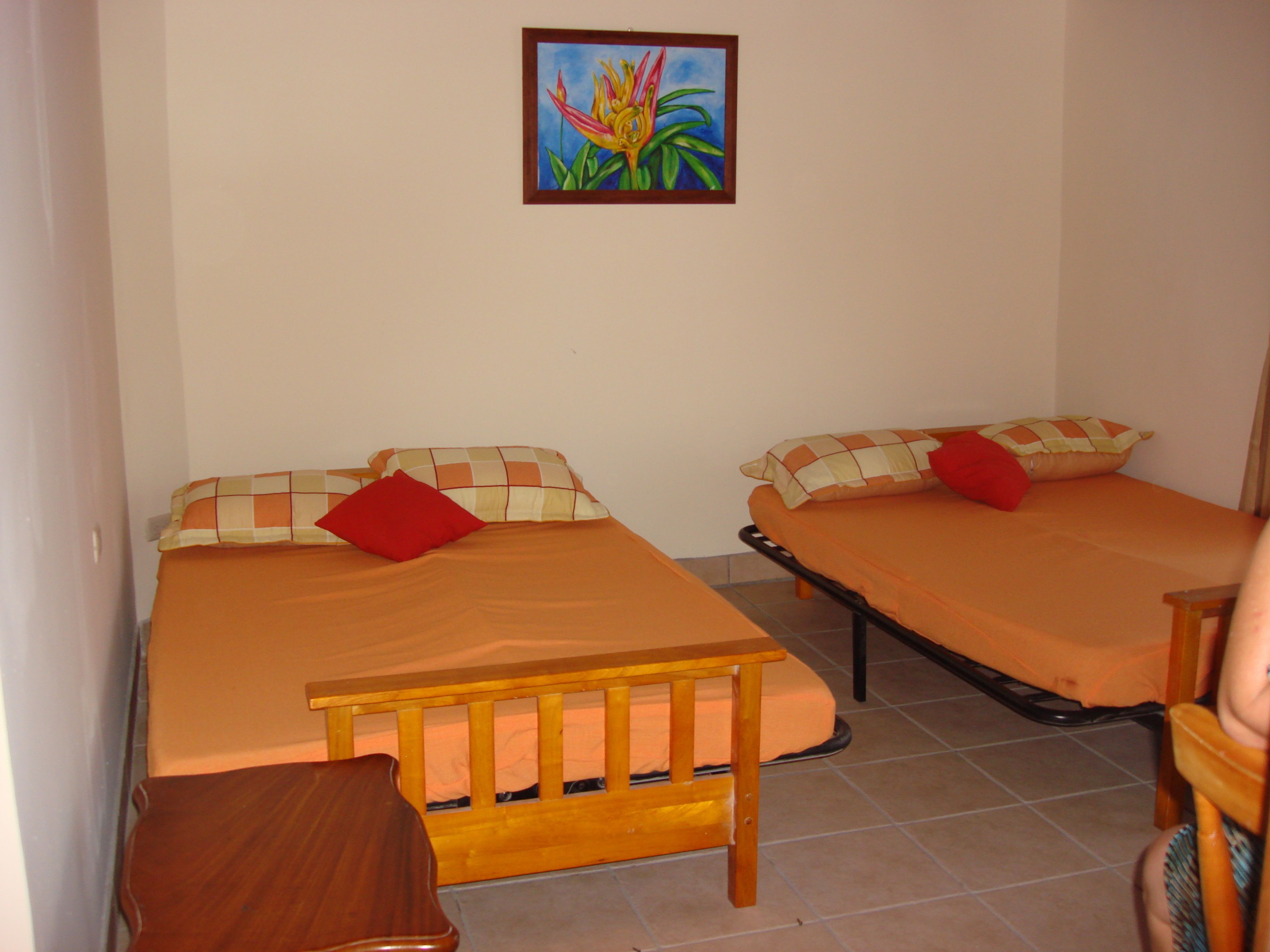 Habitación con dos camas, cada una con cabecero de madera y ropa de cama naranja. En primer plano, una mesita de madera y una silla.