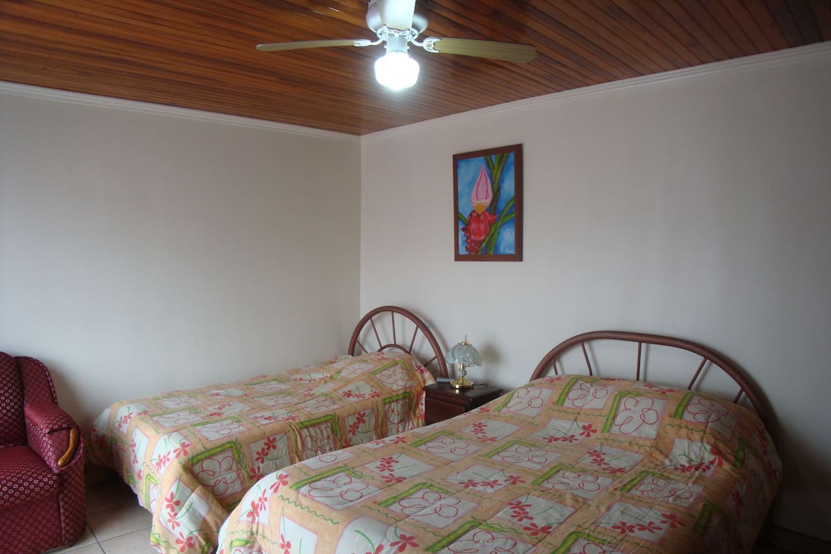 Ein Zimmer mit zwei Betten, einem Deckenventilator, einem Bild an der Wand und einem Sessel.