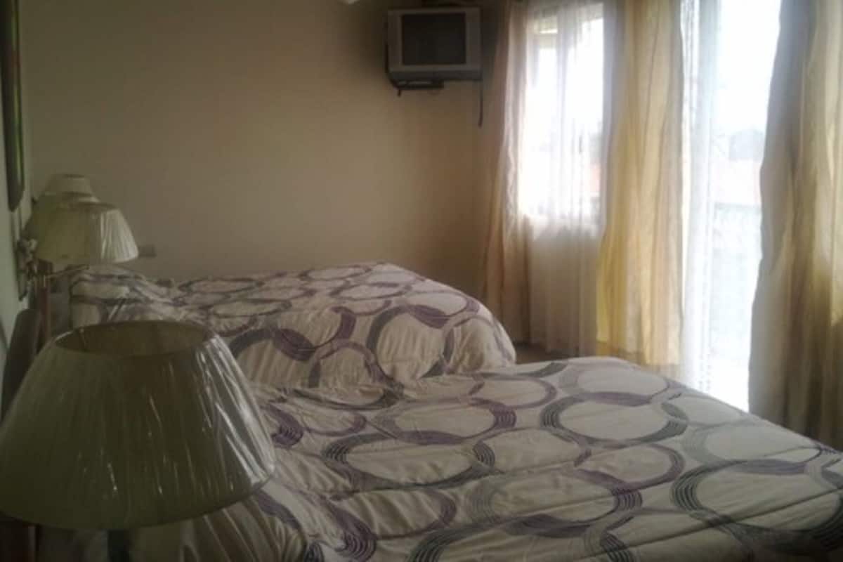 Ein Zimmer mit zwei Betten, einem Deckenventilator, einem an der Wand montierten Fernseher und einem Fenster mit Vorhängen.