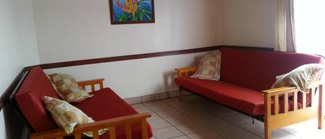 A room with two wooden sofas, one with red cushions and the other with beige cushions, a small wooden table, and a framed painting on the wall.