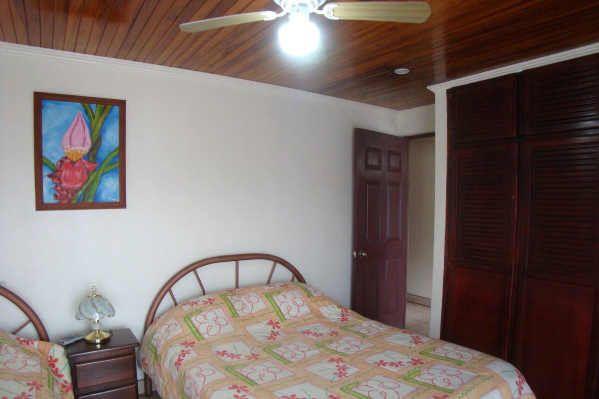 Ein Schlafzimmer mit Holzboden, einem Bett mit floraler Bettwäsche, einem Nachttisch mit Lampe und einer Tür zu einem Kleiderschrank.