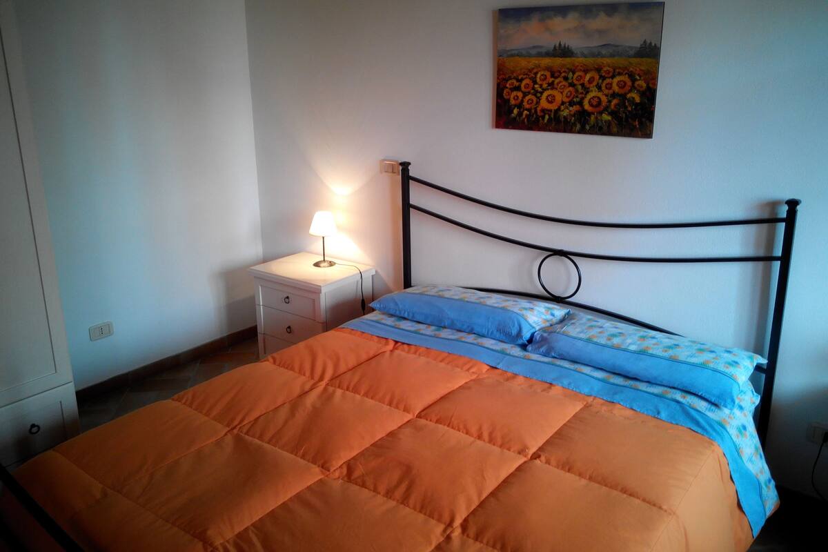Ein Schlafzimmer mit einem Bett, einem Nachttisch, einer Lampe und einem Sonnenblumenbild an der Wand.