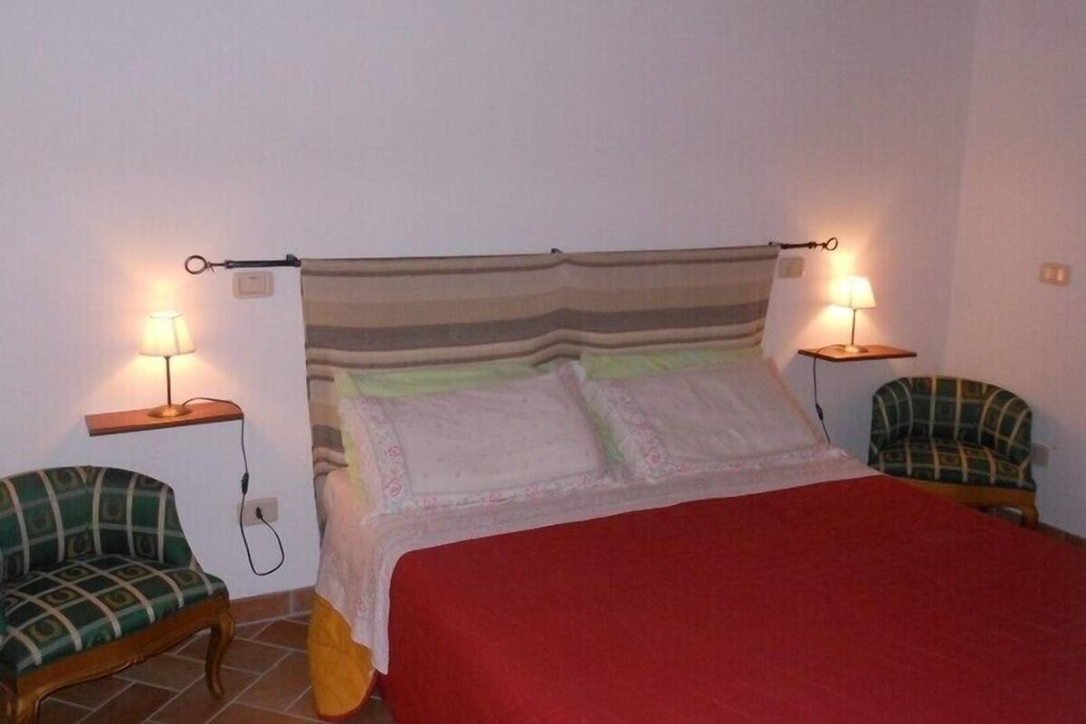 Ein Einzelbett mit roter Bettdecke, zwei Nachttische mit Lampen und zwei Polsterstühle.