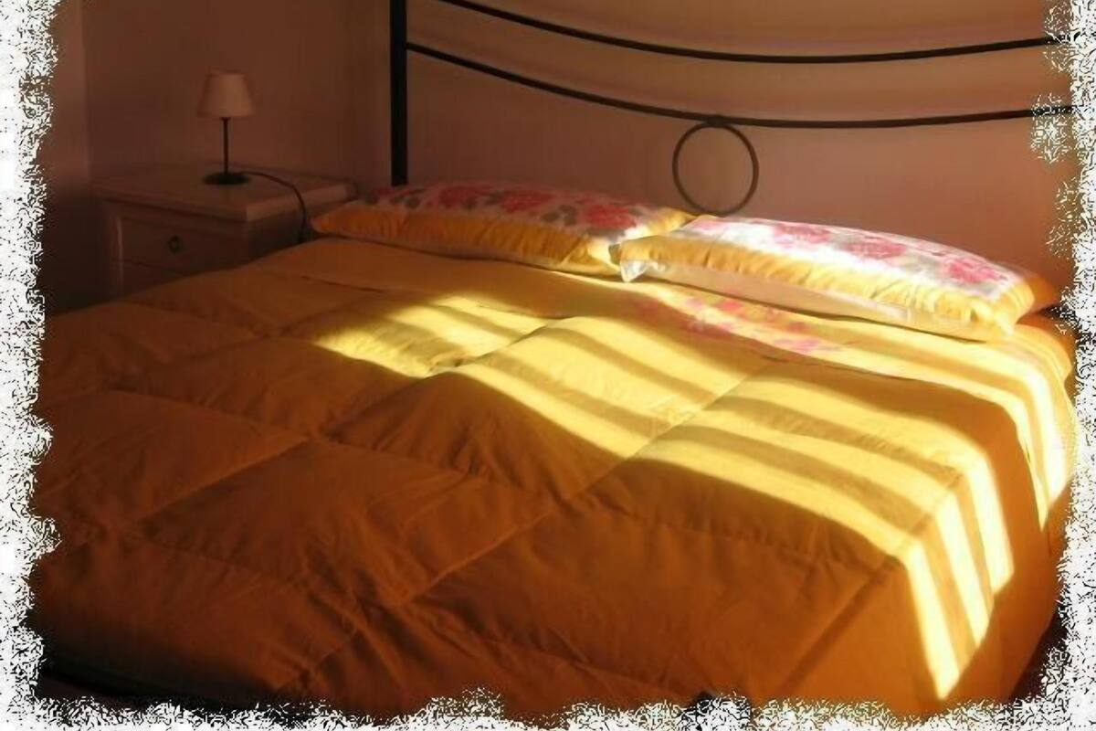 Ein Bett mit gelber Bettdecke und blumigen Kissen, ein Nachttisch mit Lampe sowie ein Kopfteil mit Rundmuster.