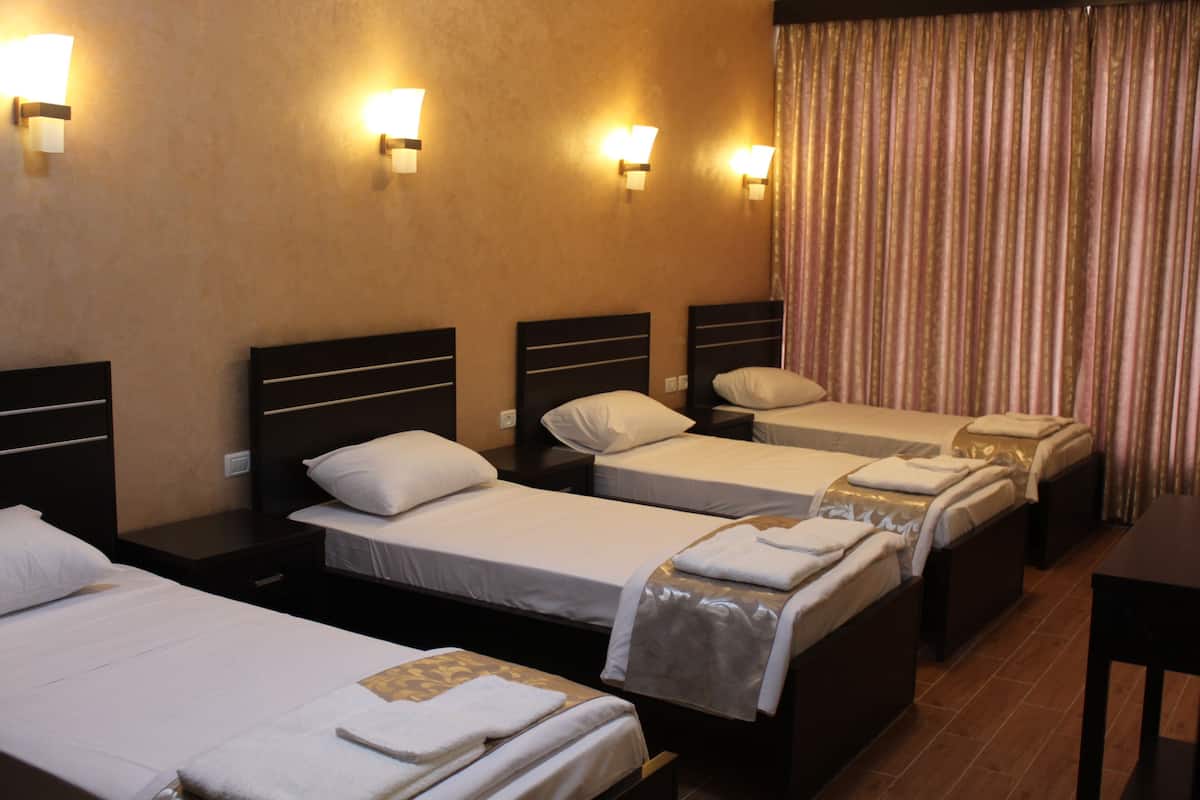A hotel room with four single beds, each with a pillow and a blanket. There are wall-mounted lights and a curtain on the window.