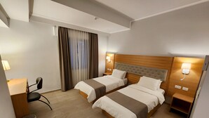 Une chambre d’hôtel avec deux lits, un bureau, une chaise, une poubelle et une fenêtre avec des rideaux.