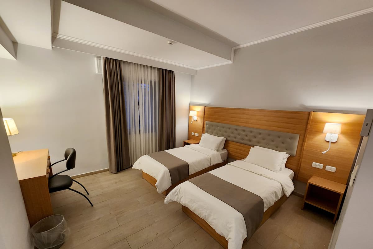 A hotel room with two beds, a desk, a chair, a waste bin, and a window with curtains.