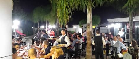 Un área de comedor al aire libre con varias mesas y sillas, rodeada de palmeras e iluminada por luces colgantes.