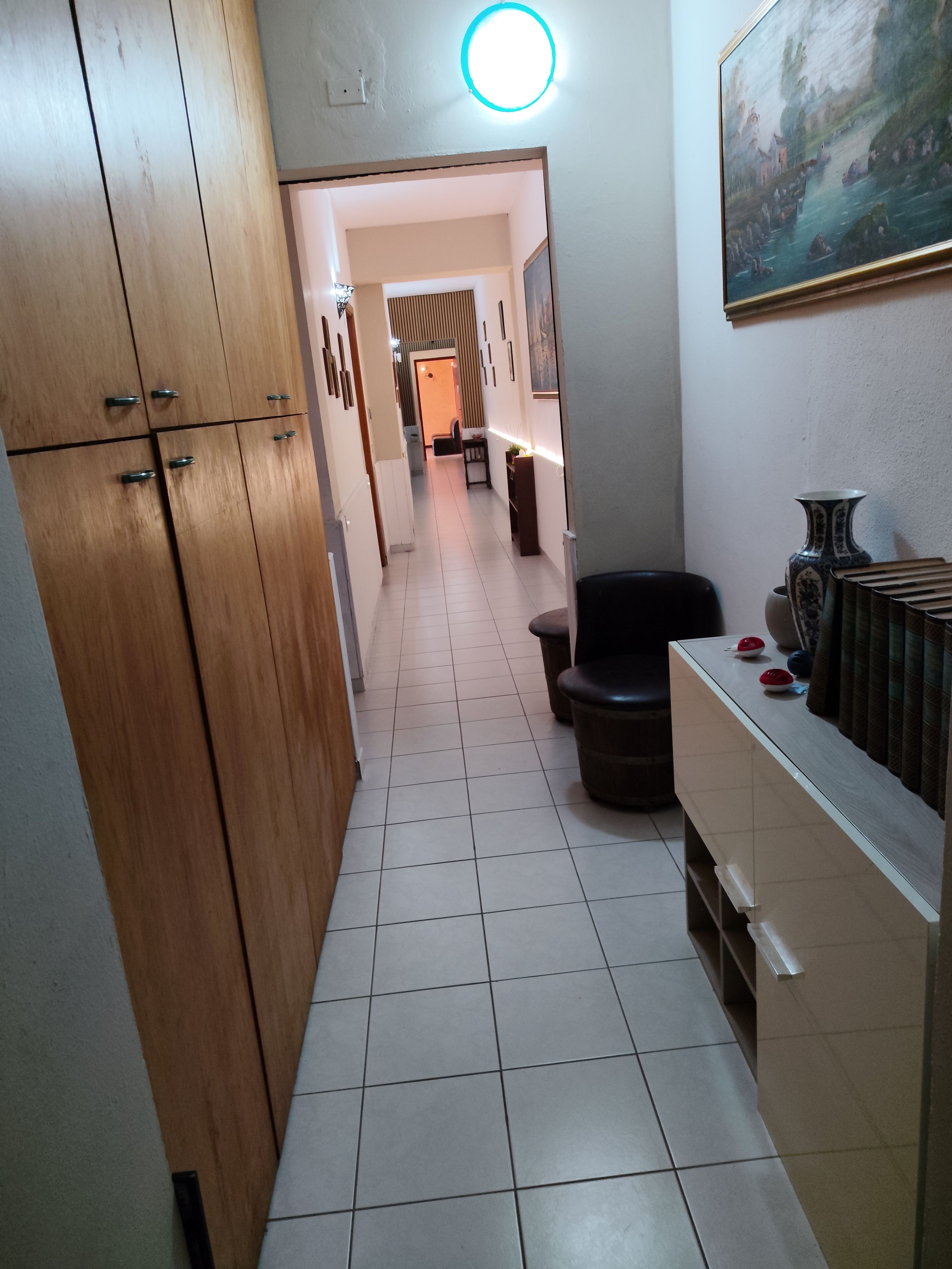 Un pasillo estrecho con un armario de madera, piso de baldosas, una silla negra y un mueble blanco con adornos.