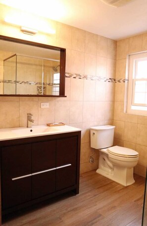A bathroom with a white sink, dark wooden cabinets, a white toilet, and a mirror with a tiled backsplash. - Paraquet Apartments (Paget Parish)