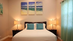 A hotel room with a large bed, two bedside lamps, and three paintings of the ocean on the wall. - Paraquet Apartments (Paget Parish)