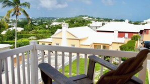 A wooden chair on a balcony overlooking a residential area with houses, a palm tree, and a clear sky. - Paraquet Apartments (Paget Parish)