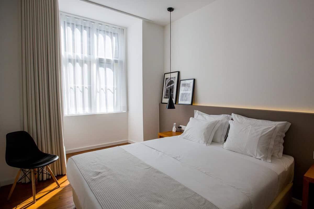 Une chambre d’hôtel moderne avec un grand lit, une chaise, une table de chevet et une fenêtre avec des rideaux légers.