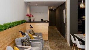 Un área de recepción con mostrador, una sala de espera con sofás grises y cojines amarillos, y suelo de baldosas.