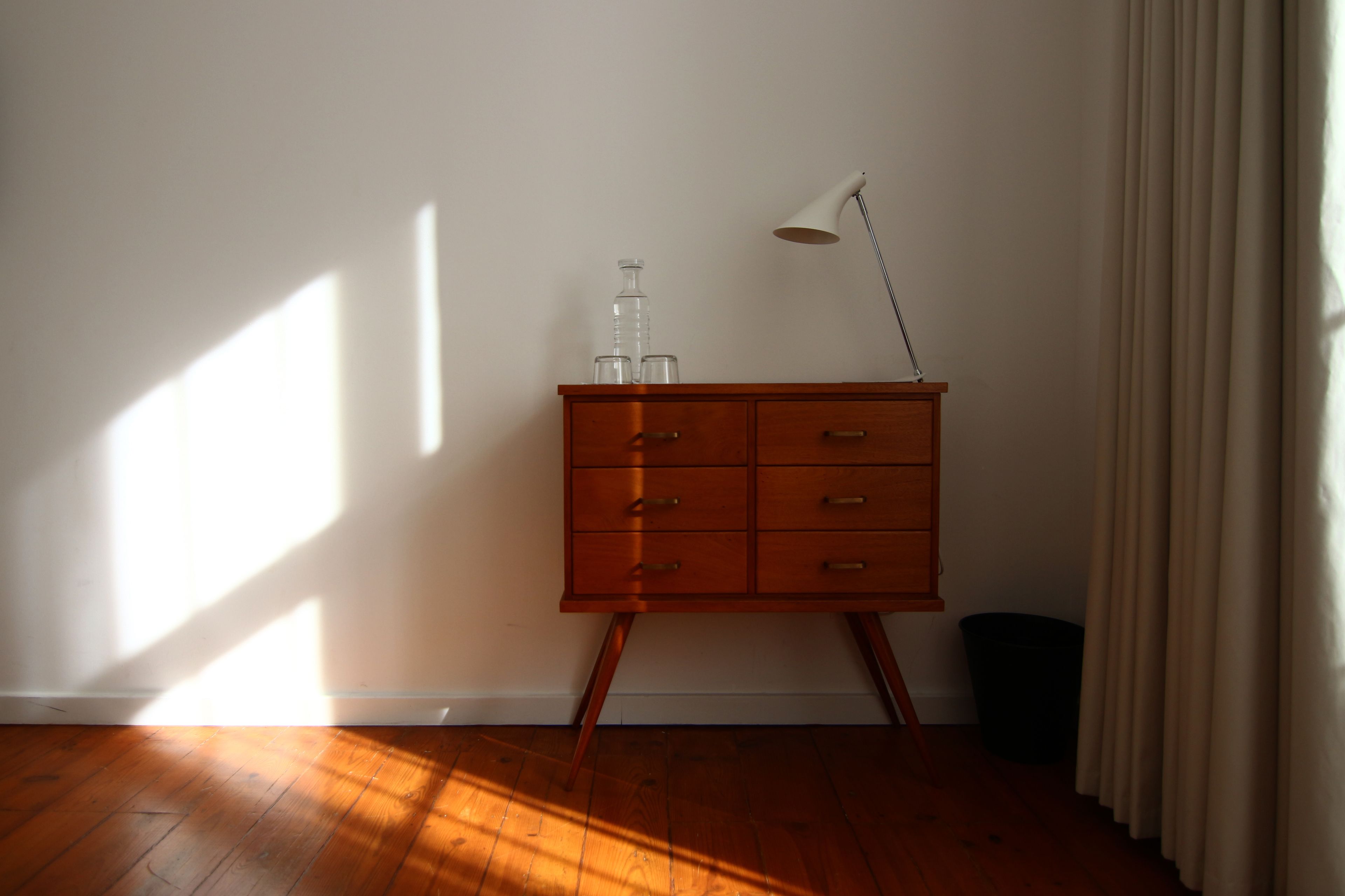 Een houten dressoir met laden, een lamp en een prullenbak in een kamer met houten vloeren en een raam waardoor schaduwen vallen.