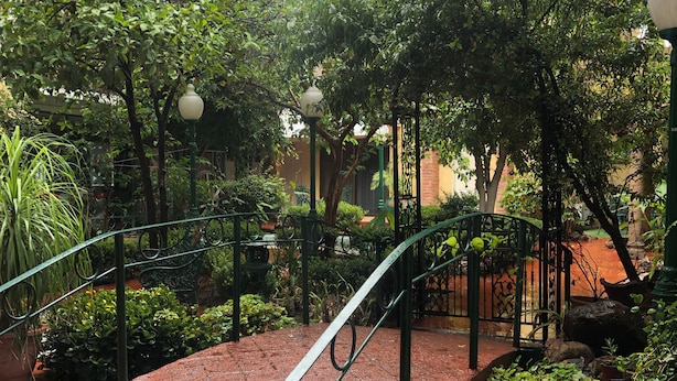 A brick pathway leading to a small bridge over a water feature, surrounded by lush greenery and a traditional building with a tiled roof. - Inn Galerias Hotel (Aguascalientes)