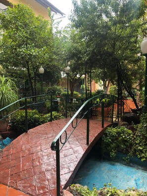 A brick pathway leading to a small bridge over a water feature, surrounded by lush greenery and a traditional building with a tiled roof. - Inn Galerias Hotel (Aguascalientes)