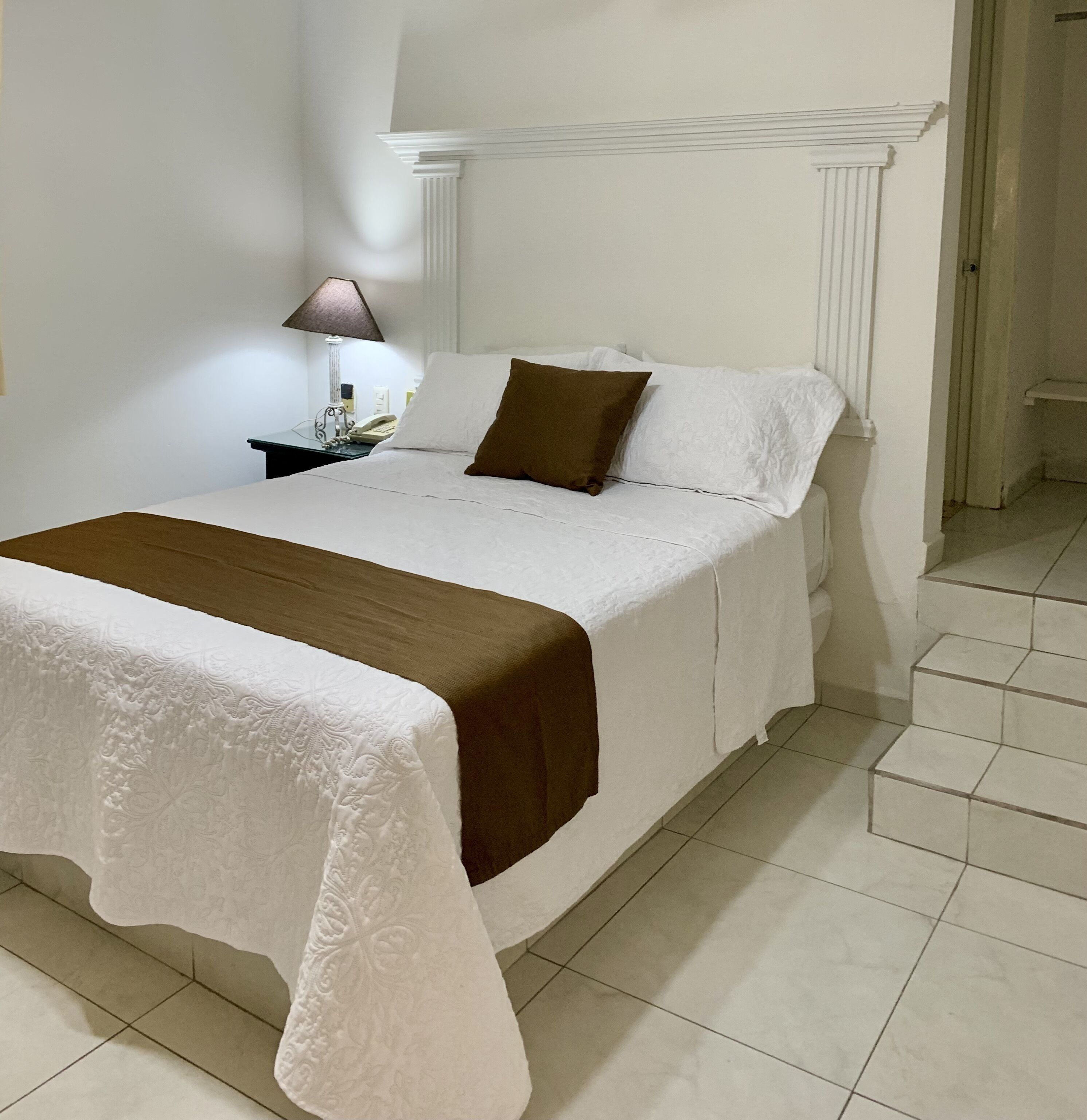 Ein ordentlich gemachtes Bett mit weißer und brauner Bettdecke, ein Nachttisch mit Lampe und eine weiße Wand mit dekorativer Leiste.