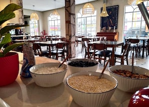A dining area with multiple tables and chairs, bowls of food, and a potted plant. - Inn Galerias Hotel (Aguascalientes)