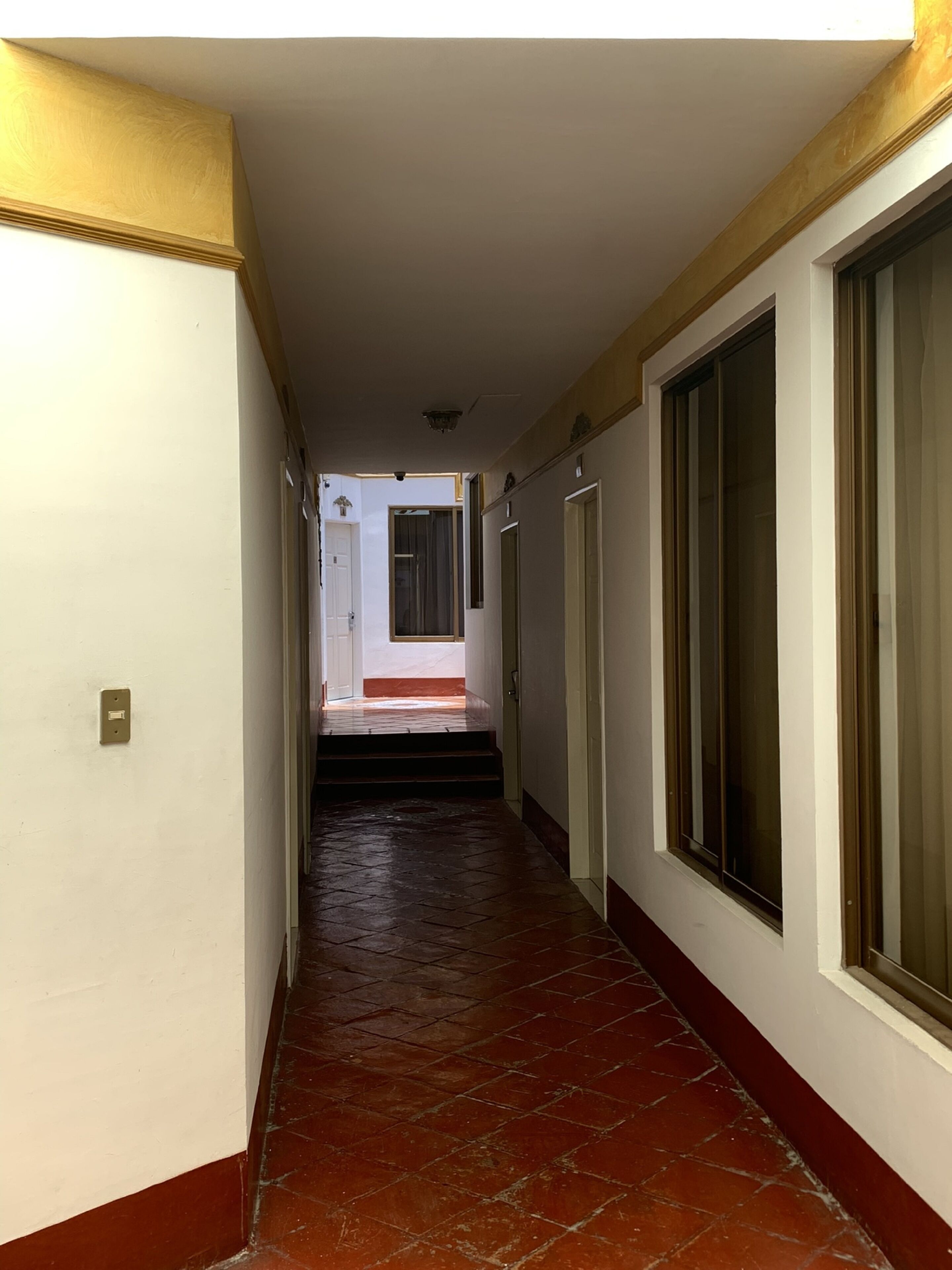 A narrow hallway with a red-tiled floor, white walls, and large windows on one side.